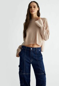 Maglione corto di colore beige chiaro con maniche lunghe, abbinato a pantaloni cargo blu scuro con tasche e dettagli di cucitura a contrasto.