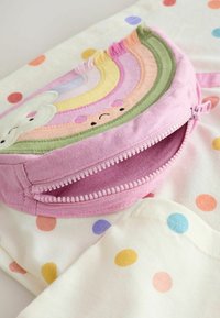 Pasteļrozā rāvējslēdzēja somiņa ar izšūtu varavīksnes un mākoņa dizainu, novietota uz krēmkrāsas auduma ar daudzkrāsainiem punktiņiem.