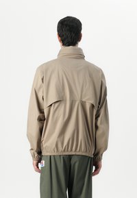Beige lettjakke med høy krage, elastisk liv og detaljer med ventiler bak. Materialet ser glatt og vannavvisende ut.