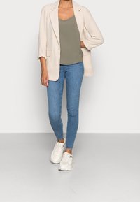 Persona che indossa un blazer a quadri beige chiaro sopra una maglia verde oliva, jeans skinny blu e sneakers bianche spesse, in piedi su un pavimento grigio.