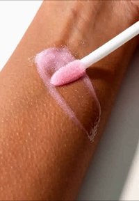 Un cotton swab applica un gel rosa lucido su un braccio abbronzato, rivelando una texture della pelle liscia sottostante. Lo sfondo è di un bianco neutro.