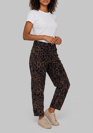 Vrouw draagt een wit topje met korte mouwen, hoge bruine broek met zwarte vlekken en beige sneakers, staand tegen een effen grijze achtergrond.