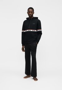 Homme portant un sweat à capuche Moschino noir avec bande logo et pantalon noir assorti, debout pieds nus devant un fond clair uni.