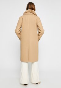Beige wollen blend jas met een nepbont kraag, rechte snit, met een enkele split aan de achterkant, gecombineerd met witte wijde broeken en hakken.