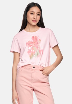 Woman wearing a light pink T-shirt with abstract pink and gold patterns, paired with high-waisted pink pants, standing with one hand in pocket.