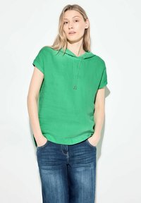 Top verde in lino a maniche corte con cappuccio e coulisse, abbinato a jeans in denim blu. Il tessuto appare leggero e testurizzato.