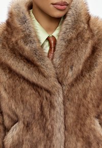 Brun faux fur frakke med en blød tekstur, bred krave og skrå frontlommer, parret med en lys grøn skjorte og en brun slips.