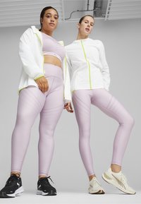 Två modeller bär matchande ljuslila leggings med perforerade mönster och koordinerande sport-bh:ar, tillsammans med en vit zip-up jacka med neonaccent.