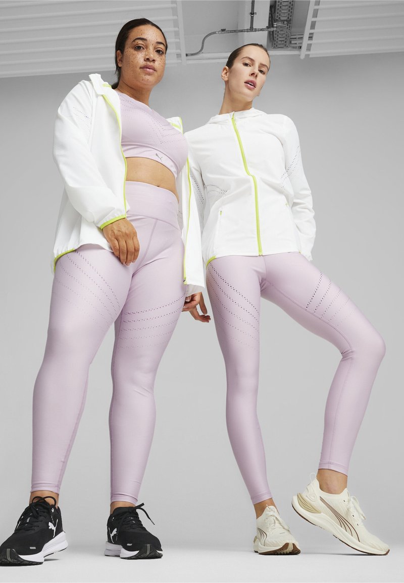 Två modeller bär matchande ljuslila leggings med perforerade mönster och koordinerande sport-bh:ar, tillsammans med en vit zip-up jacka med neonaccent.