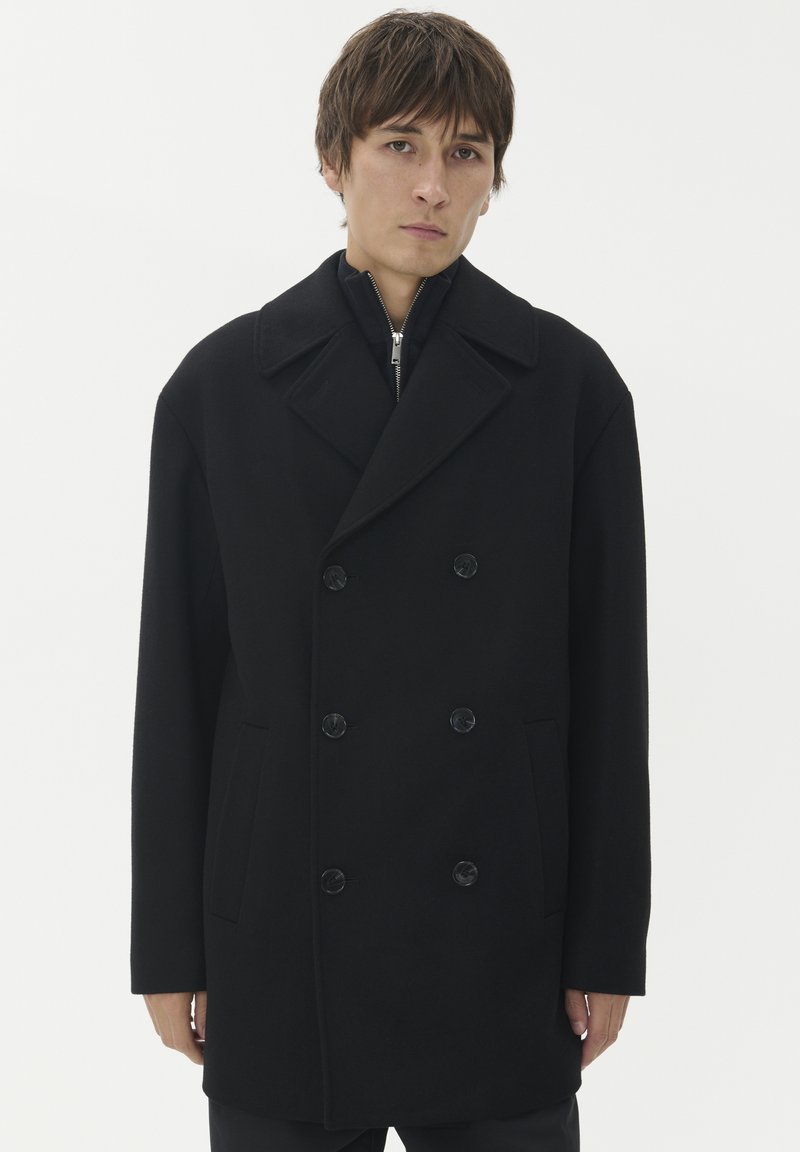 Cappotto in lana nera a doppio petto con ampi rever, due tasche frontali e bottoni rivestiti in tessuto. Materiale testurizzato, vestibilità strutturata.