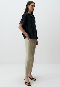 Camicetta nera a maniche corte con colletto, abbinata a pantaloni aderenti beige chiaro e ciabatte nere. L'outfit ha una silhouette rilassata.