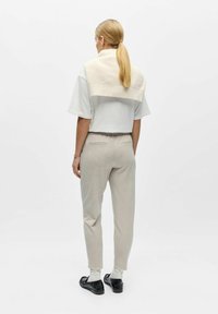 Femme aux cheveux blonds attachés en queue de cheval, portant un t-shirt blanc, un pantalon beige, un pull crème posé sur ses épaules, des chaussettes blanches et des mocassins noirs, vue de dos.