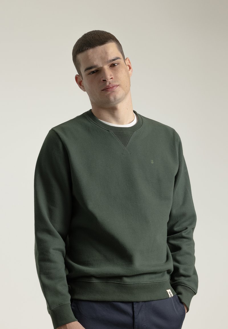 Sudadera verde de material suave, con cuello redondo, mangas largas, puños acanalados y un pequeño logo en el pecho. Capa base blanca visible.