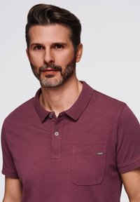 Polo shirt bordeaux con colletto, maniche corte e una tasca frontale. Il tessuto sembra morbido con una sottile texture e un dettaglio a bottone.