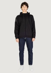 Felpa nera con zip e cappuccio regolabile, realizzata in un tessuto strutturato e con dettagli a contrasto sui lati, abbinata a jeans scuri e sneakers bianche.