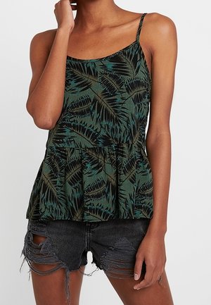 Kobieta ubrana w zielono-czarny, bezrękawnik z cienkimi ramiączkami, z wzorem liści oraz czarne, postrzępione szorty jeansowe.