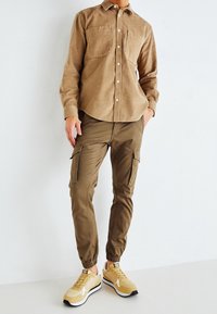 Camicia a bottoni in velluto a coste marrone con due tasche sul petto, abbinata a pantaloni cargo olivastro con tasche laterali e sneakers atletiche beige.