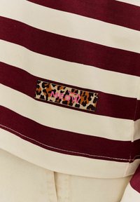 Pruhované bavlněné tričko v barvách mahagonové a krémové, s nášivkou s leopardím vzorem a nápisem "MEOW" v růžovém textu. Standardní design s kulatým výstřihem.