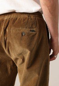 Bruine corduroy broek met een gestructureerd oppervlak, voorzien van een achterzak en een knoopsluiting. Het merklabel is zichtbaar op de zak.
