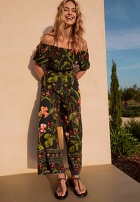 Salopetă cu umerii descoperiți din țesătură închisă la culoare, cu modele florale colorate, talie strânsă, picioare largi și cusături decorative la tiv.