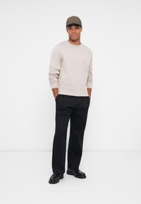 Sudadera beige con detalle bordado, pantalones negros y zapatos negros. El modelo lleva una gorra de cuadros marrón, de pie contra un fondo blanco.