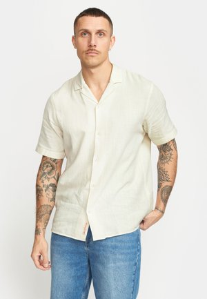 Korte mouwen, button-up shirt in lichtbeige, gemaakt van een textuurstof. Heeft een kraag en een zijlabel van het merk. Draagt met blauwe spijkerbroek.