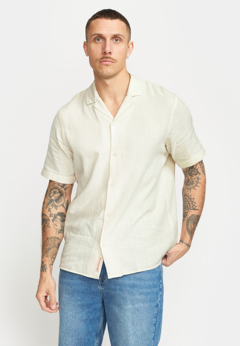 Korte mouwen, button-up shirt in lichtbeige, gemaakt van een textuurstof. Heeft een kraag en een zijlabel van het merk. Draagt met blauwe spijkerbroek.