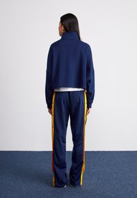 Navyfarbener, kürzerer Sweatshirt mit hohem Kragen und lockerer Passform, kombiniert mit navyfarbenen Hosen mit gelben Seitenstreifen und Druckknöpfen. Glatte Textur.