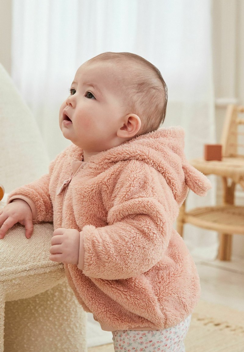 Haut à capuche en polaire couleur pêche avec une texture douce et une capuche, présentant une fermeture à l'avant et des coins arrondis. Un enfant se tient à côté d'une surface texturée.