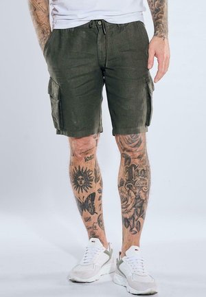 Mann trägt dunkelgrüne Cargoshorts, weiße Sneakers, und zeigt tätowierte Beine mit Sonnen-, Schmetterlings- und Blumendesigns.