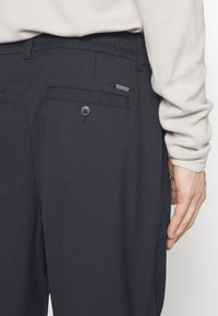 Pantaloni grigi scuri con vita elastica, tasche laterali e posteriori, chiusura con bottone e un piccolo etichetta del marchio sulla tasca. Tessuto morbido.
