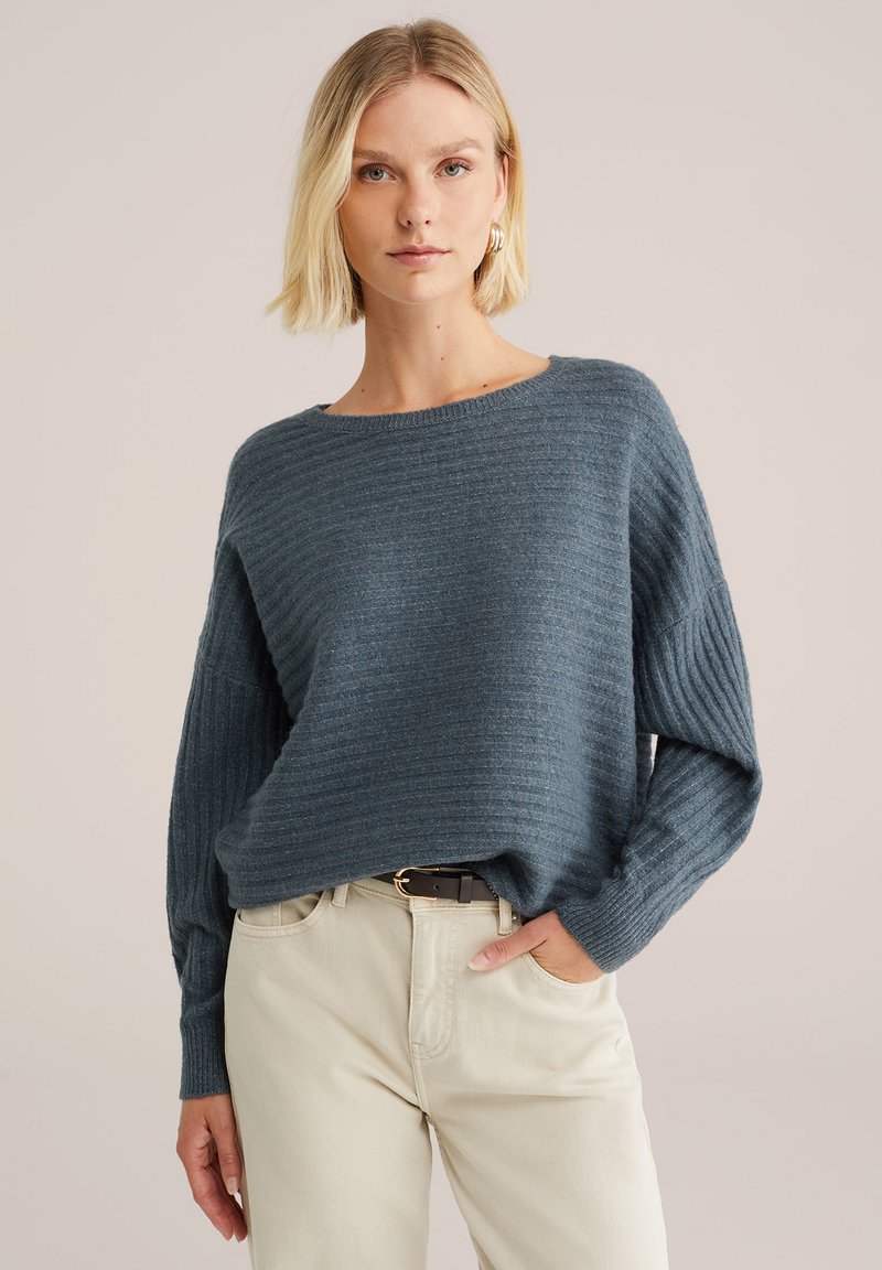 Blauer, gestreifter Pullover mit rundem Halsausschnitt und langen Ärmeln, kombiniert mit hellen Hosen. Der Stoff wirkt weich und strukturiert.