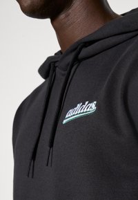 Crna dukserica s kapuljačom napravljena od glatkog materijala, s vezenim "adidas" logom u bijeloj i zelenoj boji na prsima, s vezicama za kapuljaču.