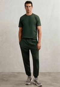 Set sportiv de culoare olive, format dintr-un tricou scurt, ajustat, cu mânecă scurtă și pantaloni jogger conici cu pliuri frontale, asortați cu teniși gri.