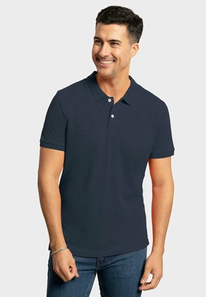 Camisa polo navy con tapeta de tres botones y un sutil logo de la marca en el pecho izquierdo, combinada con jeans de mezclilla azules. Textura suave, mangas cortas.
