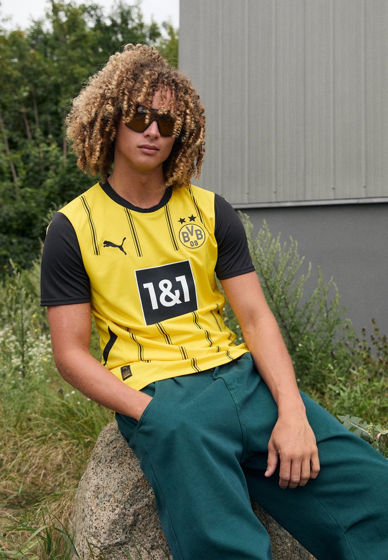 Gul og sort kortærmet sports trøje med lodrette striber og logoer, parret med grønne bukser. Model poserer på en sten, iført solbriller.