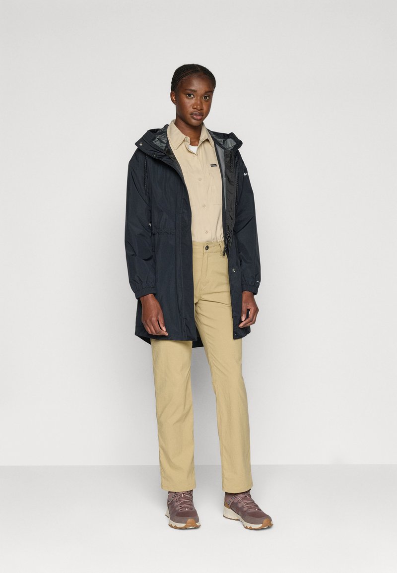 Longue veste imperméable noire avec capuche, chemise beige clair, pantalon beige et chaussures de randonnée marron avec des accents roses.