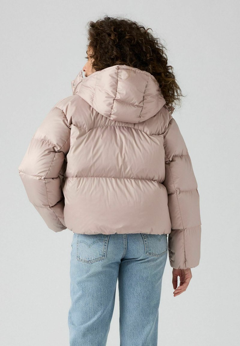 Chaqueta acolchada de color rosa claro con capucha, diseño acolchado y puños elásticos, combinada con unos vaqueros sueltos de color azul claro.