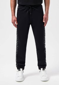 Zwarte trainingsbroek met een koord in de taille, ribgebreide manchetten en gestreepte zijstrips. Gecombineerd met witte sneakers voor een sportieve uitstraling.