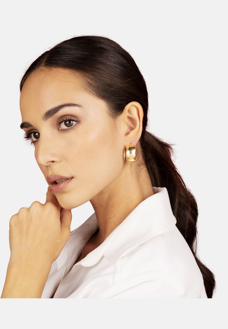 Orecchini a cerchio in oro con finitura lucida, forma circolare e leggera texture. Presentati su un modello con una coda di cavallo elegante, che indossa una camicia bianca con colletto.