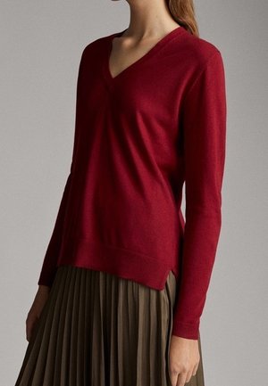 Donna che indossa un maglione aderente a scollo a V di colore rosso intenso abbinato a una gonna plissettata a vita alta verde oliva, in piedi davanti a uno sfondo semplice.