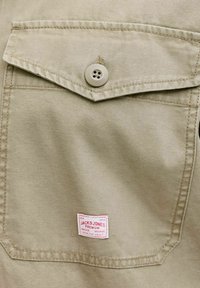 Beige Stofftasche mit Klappe, gesichert durch einen beigen Knopf, und einem kleinen weißen Etikett mit der Aufschrift "Jack & Jones Premium" nahe dem unteren Rand.