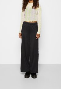 Frau in einem grünen gemusterten langärmeligen Crop-Top, schwarzen weiten Hosen mit seitlichen Streifen und schwarzen klobigen Schuhen, die vor einem weißen Hintergrund steht.