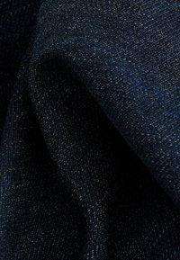 Sötétkék farmeranyag sima textúrával, amelyben finom árnyalateltérések és szoros szövés található, lágy hullámokban redőkben omlik.