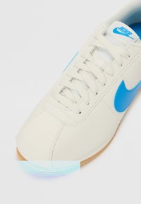 Balti tenisaki ar zilu Nike zīmolu, strukturēts materiāls, noapaļota purngala forma un plakani šņores. Apakšā gumijas zole ar redzamu protektora rakstu.