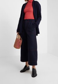 Pantalones de pierna ancha en azul marino con costuras rojas, un suéter ajustado de cuello alto rojo y un cárdigan negro. Un bolso tejido en beige y rosa. Zapatos negros con cordones.
