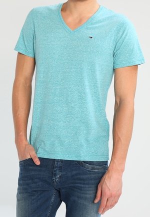 Homme portant un t-shirt bleu clair à col en V et manches courtes, ainsi qu'un jean bleu foncé, une main dans la poche.