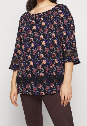 Femme portant un chemisier noir avec un imprimé floral orange et violet, manches trois-quarts avec inserts en dentelle, associé à un pantalon marron foncé.