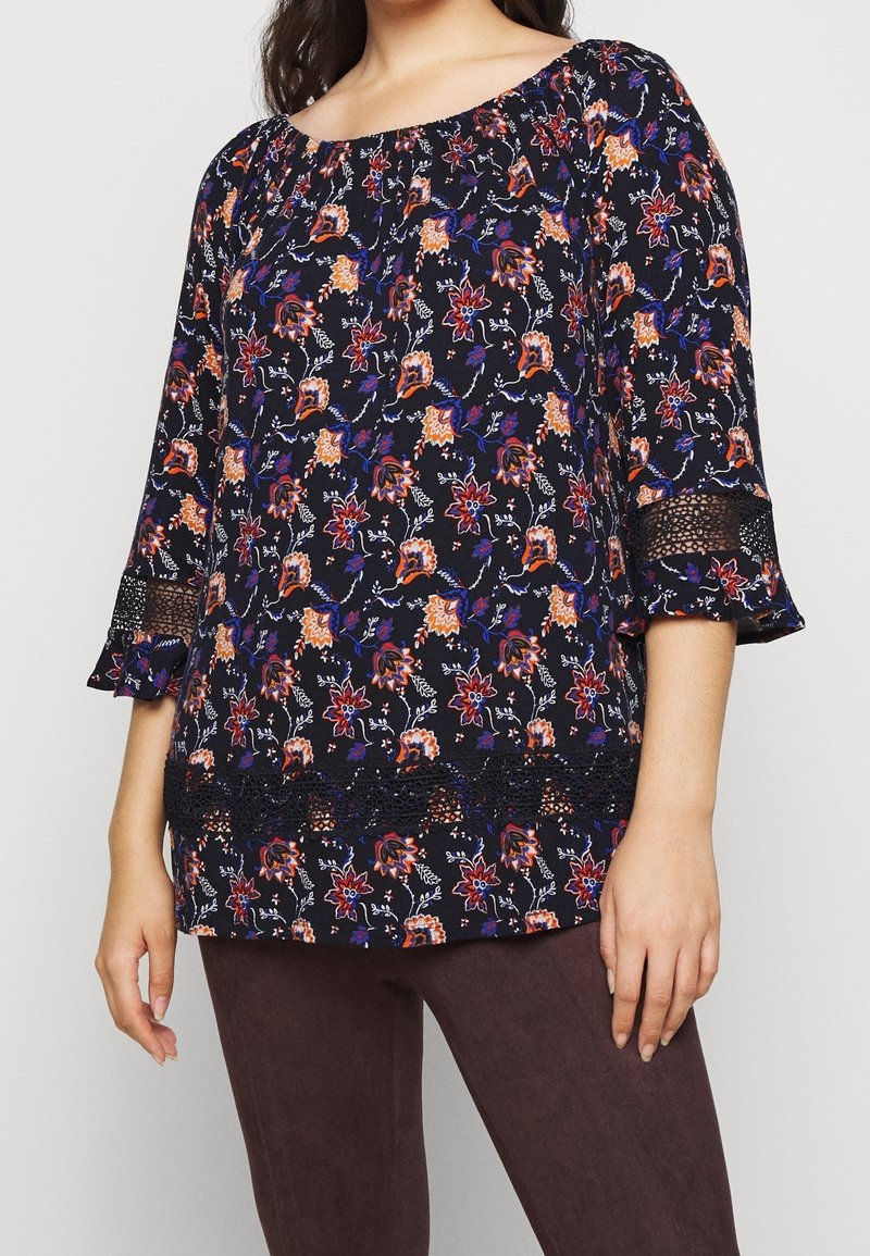 Femme portant un chemisier noir avec un imprimé floral orange et violet, manches trois-quarts avec inserts en dentelle, associé à un pantalon marron foncé.