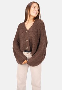 Brauner Strickcardigan mit V-Ausschnitt, übergroßen Ärmeln und drei dekorativen Knöpfen. Getragen über einem dunklen Oberteil und kombiniert mit hellen Hosen.